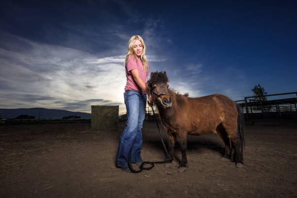 Portrait of a woman posing in a paddock on a Utah ranch, holding onto the reins of an adorable miniature horse. 